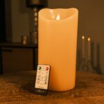 Eine wiederaufladbare LED-Wachskerze D12,5 X H25 CM steht auf einem Holztisch mit der Fernbedienung davor, während im Hintergrund sanft beleuchtete, verschwommene Möbel und Dekorationen erscheinen.
