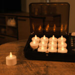 Ein Set von 12 warmweißen, wasserdichten, wiederaufladbaren LED-Wachs-/Teelichtern (Brenndauer 175h) mit Fernbedienung - eines auf einem Holztisch, andere in der schwarzen Ladestation - leuchtet sanft im Raum.
