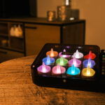 Twaalf RGB LED-was/theelichtjes (oplaadbaar, waterdicht) met afstandsbediening creëren een gezellige sfeer op een houten tafel bij een kast, hun kleurrijke lichtjes gloeien op de achtergrond.