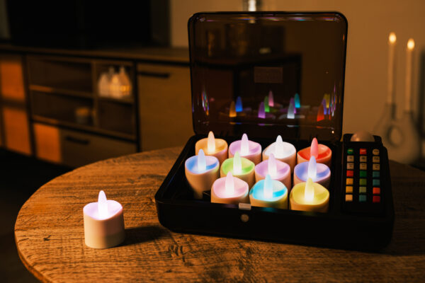 Een set van 12 RGB LED-Waxine/theelichtjes oplaadbaar waterdicht kaarsen met afstandsbediening staan in een open zwarte kist op een ronde houten tafel, één buiten de kist geplaatst; een moderne woonkamer is op de achtergrond.