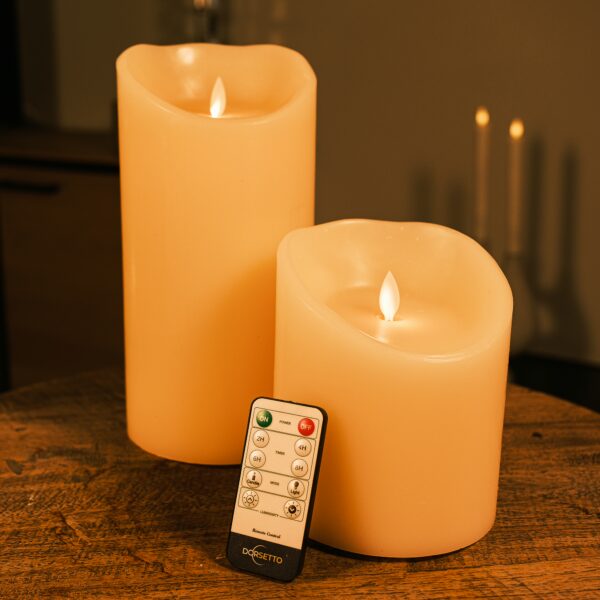 De LED wax kaarsen set 2 maten warm wit oplaadbaar DORSETTO staat op een houten tafel en gloeit met levensechte vlammen. Een afstandsbediening leunt tegen de kortere kaars en een zacht verlichte achtergrond versterkt de gezellige sfeer.