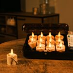 Ein schwarzer Kasten auf einem Holztisch enthält mehrere brennende LED-Kerzen und eine Fernbedienung. Eine ähnliche LED-Kerze befindet sich außerhalb des Kastens. Der Hintergrund ist sanft beleuchtet, und es sind Möbel und weitere Kerzen zu sehen.