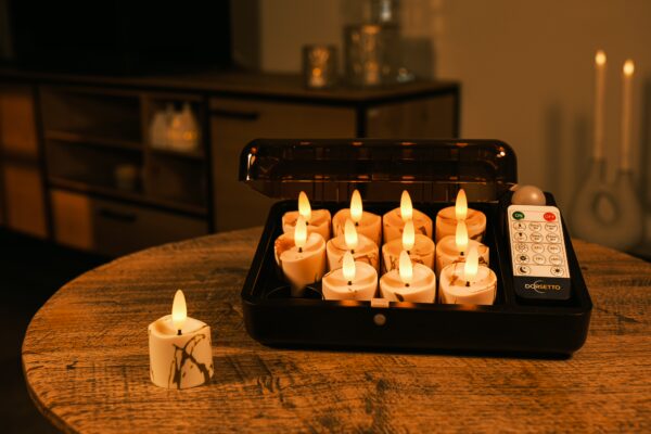 Ein schwarzer Kasten auf einem Holztisch enthält mehrere brennende LED-Kerzen und eine Fernbedienung. Eine ähnliche LED-Kerze befindet sich außerhalb des Kastens. Der Hintergrund ist sanft beleuchtet, und es sind Möbel und weitere Kerzen zu sehen.