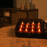 Auf einem Holztisch in einem warm beleuchteten Wohnzimmer steht eine schwarze Schachtel mit mehreren kleinen, batteriebetriebenen Kerzen ohne Flamme und einer Fernbedienung.