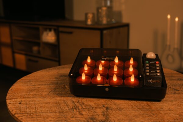 Auf einem Holztisch in einem warm beleuchteten Wohnzimmer steht eine schwarze Schachtel mit mehreren kleinen, batteriebetriebenen Kerzen ohne Flamme und einer Fernbedienung.