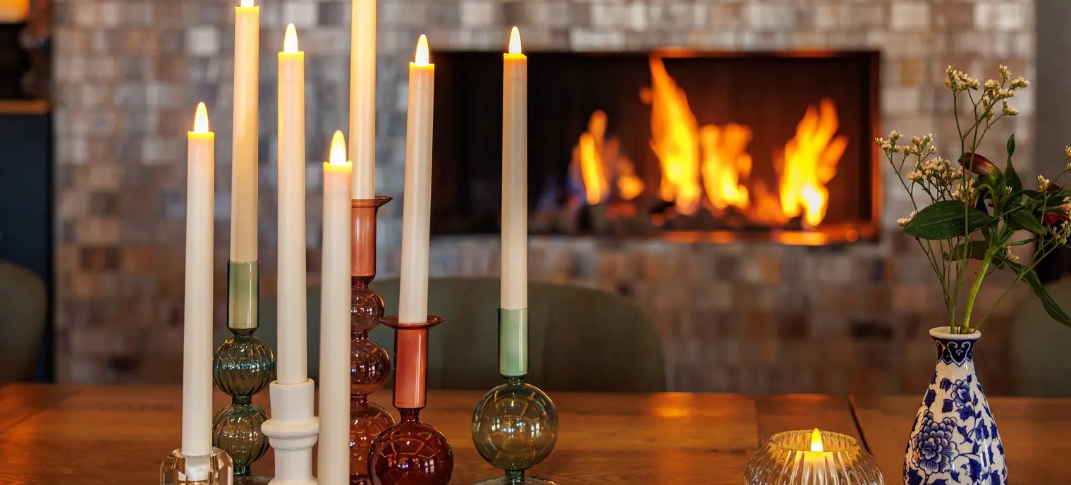 De voordelen van oplaadbare waxinelichtjes in uw huis 3 Een groep oplaadbare waxinelichtjes in kleurrijke glazen houders staat op een houten tafel, met een blauw-witte vaas bloemen in de buurt, voor een gezellige open haard met vlammen.