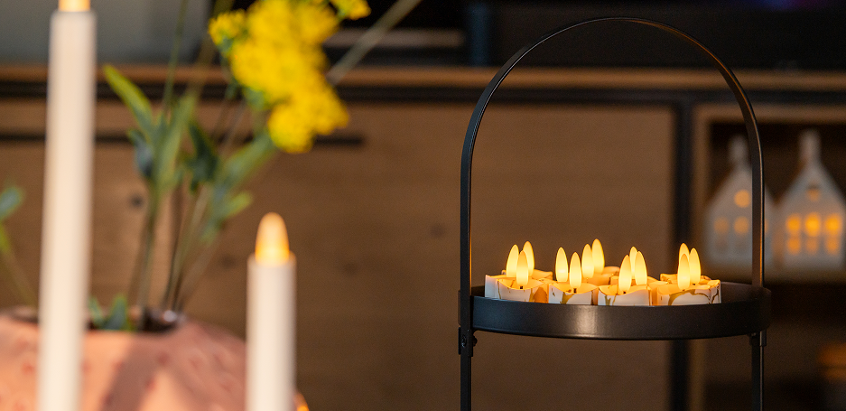 Een zwarte metalen houder toont verschillende oplaadbare waxinelichtjes die warm oplichten, met vage gele bloemen en decor op de achtergrond.