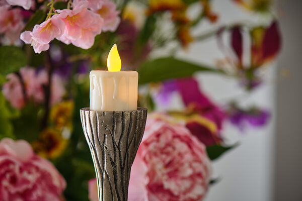 Zes oplaadbare LED-theelichtjes met kaarsvetlook, gele gloeiende vlammen en sierlijke zilveren houders voegen charme toe aan elke kamer; levendige roze en paarse bloemen creëren een zacht wazige achtergrond.