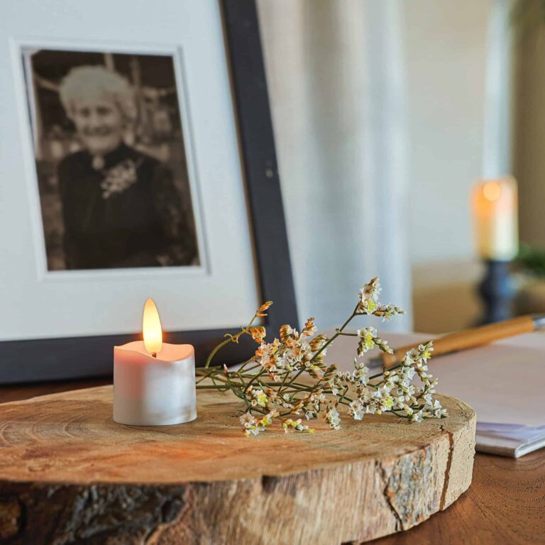 Een 3D oplaadbare led waxinelichtje (175 uur, met afstandsbediening, timerfunctie, 6 stuks), witte bloemen en een ingelijste zwart-witfoto van een oudere vrouw staan op een houten plank. Op de achtergrond zijn een kaars en een open boek zichtbaar.