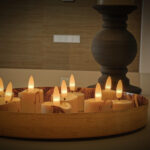 Een decoratief houten dienblad bevat zes 3D oplaadbare LED-Waxine/theelichtjes (175 uur, met afstandsbediening en timerfunctie), waarvan de gloeiende LED-vlammen een gezellige sfeer creëren met hoge kandelaars op de achtergrond.
