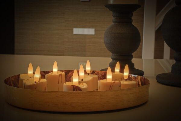 Een decoratief houten dienblad bevat zes 3D oplaadbare LED-Waxine/theelichtjes (175 uur, met afstandsbediening en timerfunctie), waarvan de gloeiende LED-vlammen een gezellige sfeer creëren met hoge kandelaars op de achtergrond.