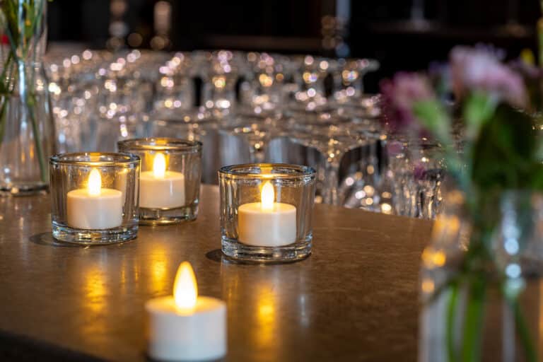 Vier LED waxine lichtjes oplaadbaar met afstandsbediening waterdicht brandtijd 175 uur flikkeren op een aanrecht en werpen warm licht, terwijl leeg glaswerk en vage bloemen op de achtergrond een gezellige, elegante sfeer creëren.