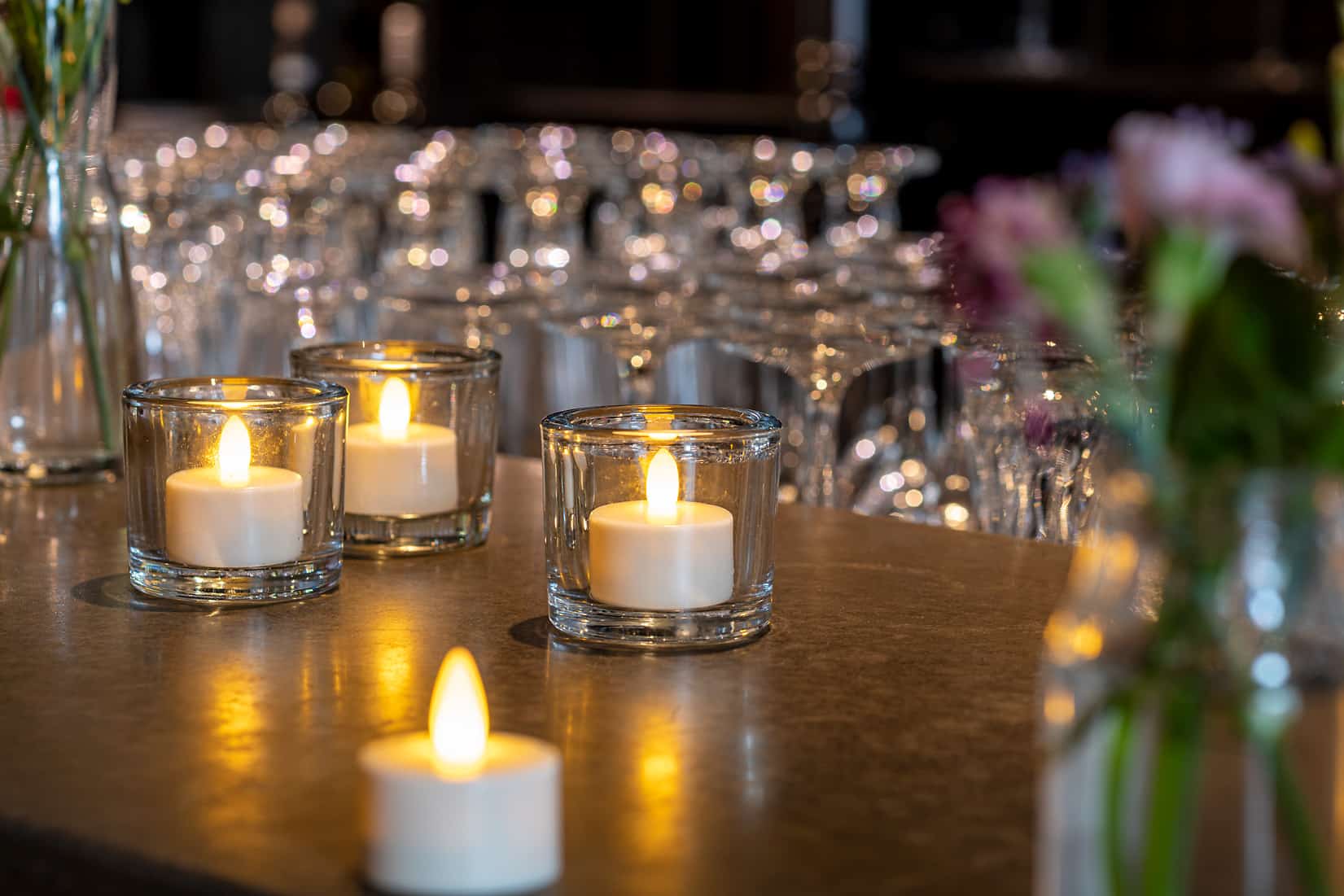 Twaalf LED-Waxine/theelichtjes waterdicht oplaadbaar brandtijd 175 uur warm wit met afstandsbediening staan in heldere houders op een aanrecht, met onscherp glaswerk en bloemen op de achtergrond, waardoor een warme, uitnodigende sfeer wordt gecreëerd.