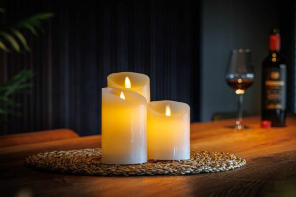 De LED wax kaarsen set 3 kaarsen DORSETTO met een warme gele gloed staat op een geweven placemat op een houten tafel. Op de zacht vervaagde achtergrond staan een glas rode wijn en een fles voor een gezellige, ontspannen sfeer.