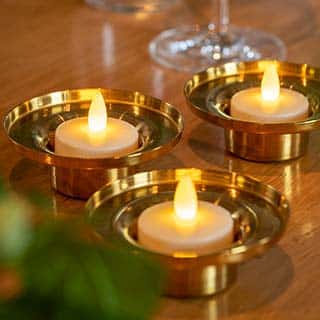 Twaalf LED-Waxinelichtjes, waterdicht en oplaadbaar met afstandsbediening (brandtijd 175 uur, warm wit), creëren een warme sfeer op hout in glanzende goudkleurige houders; op de achtergrond staan een groene plant en wijnglas.