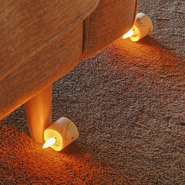 Twee 3D oplaadbare LED-Waxine/theelichtjes (175 uur, met afstandsbediening, timerfunctie) worden onder de poten van een bruine bank op een tapijtvloer geplaatst en werpen een warme oranje gloed rond de basis.