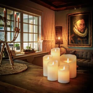 Een gezellige woonkamer heeft gloeiende kaarsen op een houten tafel, waaronder de LED wax kaars oplaadbaar DORSETTO D 7,5 X 20 CM, een bank en een groot schilderij van een man in historische kleding, allemaal warm verlicht door sfeerverlichting.