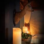 De Oplaadbare led kaars D 7,5 X 15 CM - warm wit voegt een zacht, gloeiend licht toe aan een schoorsteenmantel met groen en een hertenkop, waardoor een warme en gezellige sfeer wordt gecreëerd.
