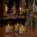 Een restauranttafel is gedekt met lege glazen, bloemen en drie DORSETTO wiederaufladbare LED-Teelichter (hohes Modell, warmweiß, 58-68 Std., mit Fernbedienung) in Glashaltern - voor een warme en elegante Atmosphäre.