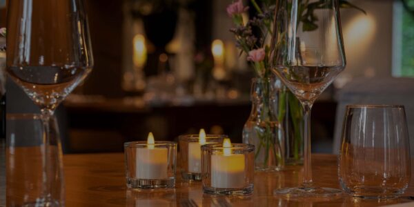 Een restauranttafel is gedekt met lege glazen, bloemen en drie DORSETTO wiederaufladbare LED-Teelichter (hohes Modell, warmweiß, 58-68 Std., mit Fernbedienung) in Glashaltern - voor een warme en elegante Atmosphäre.