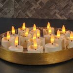 Een rond, goudkleurig dienblad bevat verschillende LED waxinelichtjes 3D (set van 12, oplaadbaar, timerfunctie, afstandsbediening) met geeloranje vlammen, die een warme sfeer creëren op een donker aanrecht tegen een bruin betegelde muur.