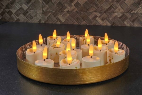 Een rond, goudkleurig dienblad bevat verschillende LED waxinelichtjes 3D (set van 12, oplaadbaar, timerfunctie, afstandsbediening) met geeloranje vlammen, die een warme sfeer creëren op een donker aanrecht tegen een bruin betegelde muur.