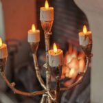 Een gouden kandelaar met vijf 3D oplaadbare LED-waxine/theelichtjes (set van 12, timerfunctie, afstandsbediening) staat voor een open haard. Het takvormige ontwerp en de gloeiende lampjes creëren een gezellige, warme sfeer.