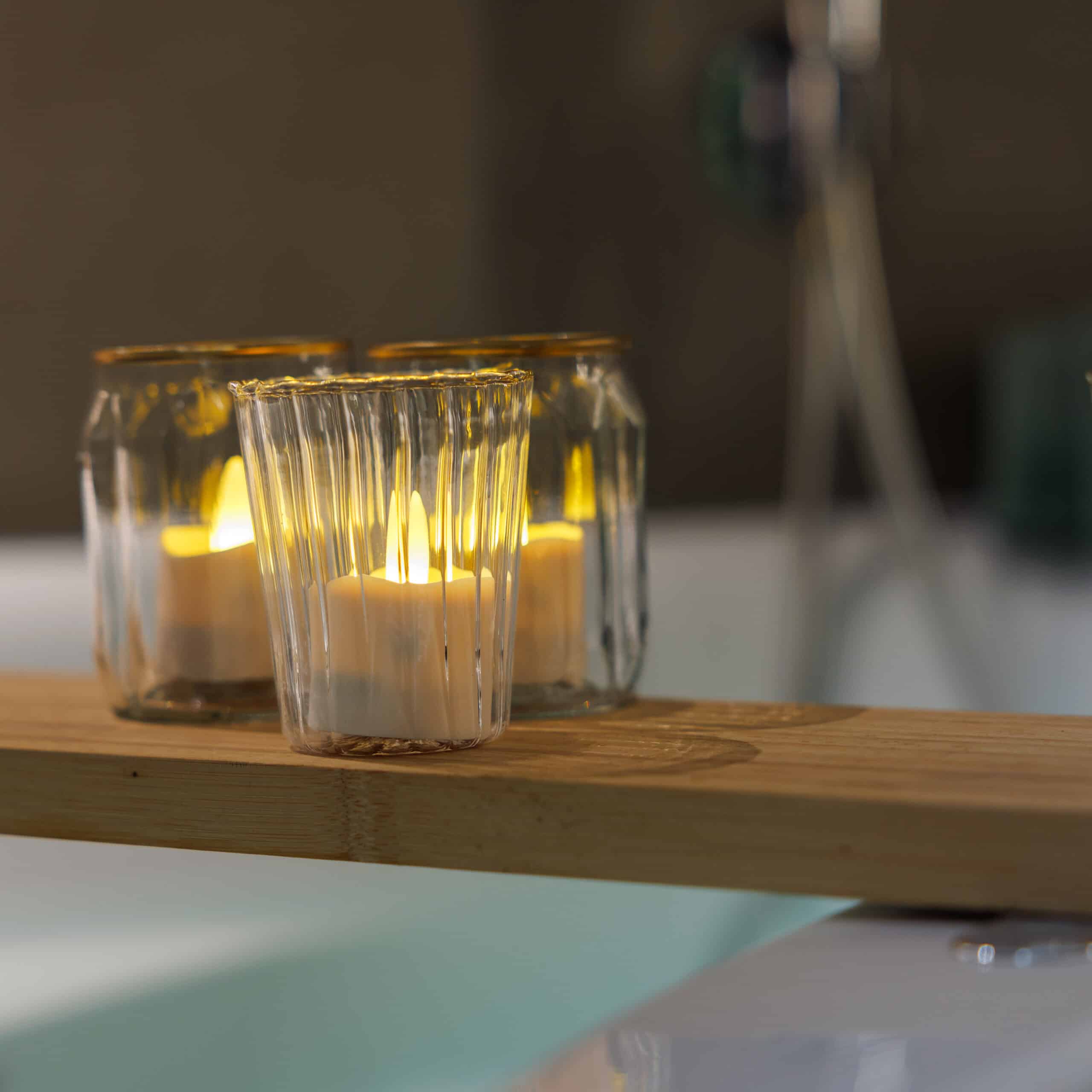 Twaalf waterdichte oplaadbare led waxinelichtjes brandtijd 175 uur, warm wit met afstandsbediening, staan in glazen houders op een houten badplank boven het bad en creëren een warme, ontspannen sfeer. De achtergrond is zacht vervaagd.