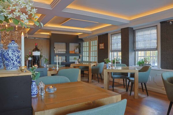Modern restaurantinterieur met houten tafels, groene stoelen met kussens, grote ramen met vouwgordijnen, zachte plafondverlichting, potplanten, decoratieve vazen en LED waxinelichtjes 3D (175 uur brandtijd, oplaadbaar) voor een gezellige sfeer.