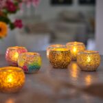 Mehrere Teelichthalter Dorsetto Mosaik Braun-Beige mit brennenden Kerzen sind auf einer spiegelnden Fläche angeordnet und verbreiten ein warmes, gemütliches Licht. Helle Blumen erscheinen sanft verschwommen im Hintergrund.