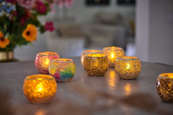 Mehrere Teelichthalter Dorsetto Mosaik Braun-Beige mit brennenden Kerzen sind auf einer spiegelnden Fläche angeordnet und verbreiten ein warmes, gemütliches Licht. Helle Blumen erscheinen sanft verschwommen im Hintergrund.