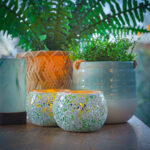 Zwei Glasmosaik-Kerzenhalter mit LED-Teelichtern wiederaufladbar mit Fernbedienung sitzen auf einem Tisch zwischen grünen Topfpflanzen in Keramik und geflochtenen Körben und schaffen ein gemütliches, natürliches Ambiente - und bis zu 400 Stunden Batterielebensdauer.