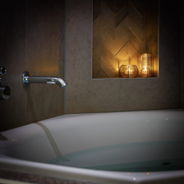 Eine moderne Badewanne mit Wasser sitzt an einer gefliesten Wand. Auf einem eingelassenen Regal darüber befinden sich zwei beleuchtete LED-Teelichter wiederaufladbar mit Fernbedienung - wasserdicht, die bis zu 400 Stunden lang ein warmes und entspannendes Ambiente schaffen.