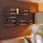 Ein modernes Wandweinregal mit mehreren Fächern fasst mehrere Weinflaschen und die LED-Wachskerze wiederaufladbar DORSETTO D 7,5 × 20 CM, die vor einer gemusterten braunen Wand in einem stilvollen, warm beleuchteten Raum steht.