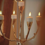 Sechs wiederaufladbare LED-Teelichter (3D) mit bis zu 400 Stunden Brenndauer, Fernbedienung und Timerfunktion sind in einem Innenraum in warmem Licht angeordnet; im Hintergrund erscheint ein verschwommener brauner Stuhl.