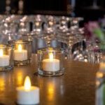 Vier LED-Teelichter wiederaufladbar mit Fernbedienung und bis zu 400 Stunden Brenndauer sind auf einem Tisch arrangiert. Im Hintergrund sorgen verschwommene Gläser und Blumenvasen für eine warme, einladende Atmosphäre.