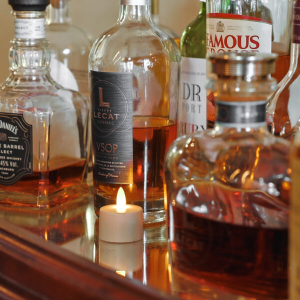 Mehrere Spirituosenflaschen, darunter Cognac und Whiskey, stehen auf einer spiegelnden Fläche neben einem LED-Teelichter wiederaufladbar mit Fernbedienung - wasserdicht - bis zu 400 Stunden Brenndauer.