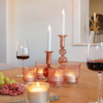 Ein Holztisch mit wiederaufladbaren LED-Teelichtern (12 Stück, warmweiß), die in rosafarbenen Glashaltern neben Wein und Wurstwaren leuchten, schafft eine gemütliche, elegante Atmosphäre; jedes Teelicht hält bis zu 400 Stunden und lässt sich einfach per Fernbedienung steuern.