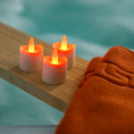 Zwölf wiederaufladbare RGB-LED-Teelichter mit bis zu 140 Stunden Brenndauer und Fernbedienung leuchten auf einem Holzbrett neben einem orangefarbenen Handtuch, vor unscharfem, blauem Hintergrund für Spa-Atmosphäre.