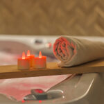 Ein aufgerolltes beiges Handtuch und drei RGB-LED-Teelichter (wiederaufladbar, wasserdicht, mit Fernbedienung) sitzen auf einer Holzplanke über einem Whirlpool und schaffen ein entspannendes Spa-Ambiente.