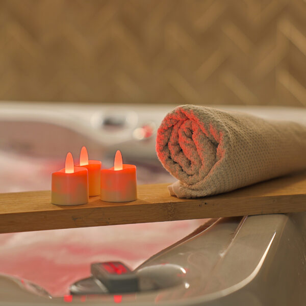 Ein aufgerolltes beiges Handtuch und drei RGB-LED-Teelichter (wiederaufladbar, wasserdicht, mit Fernbedienung) sitzen auf einer Holzplanke über einem Whirlpool und schaffen ein entspannendes Spa-Ambiente.