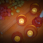 Eine gemütliche Szene mit drei warmweißen wiederaufladbaren LED-Teelichtern (bis 400h, 12er-Pack, Fernbedienung), Braunglashaltern, Käse, Salami, Weintrauben auf einem Brett und einem Glas Rotwein auf einem Holztisch.