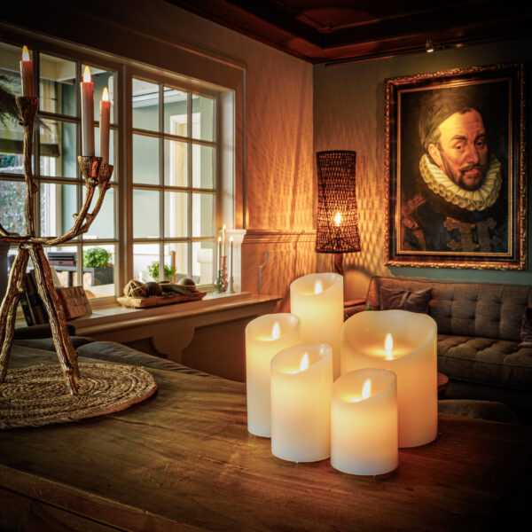 In einem gemütlichen Wohnzimmer leuchtet die LED-Wachskerze wiederaufladbar DORSETTO (7,5 × 20 CM) auf einem Holztisch, sanfte Beleuchtung, ein Kandelaber, ein bequemes Sofa und ein großes Porträt eines Mannes in historischer Kleidung an der Wand.