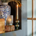 Eine blau-weiße Deko-Vase mit getrocknetem Gras, ein LED-Teelicht wiederaufladbar mit Fernbedienung und ein kleiner Holzklotz stehen auf einem dunklen Regal neben einem Milchglasfenster mit Holzumrandung.