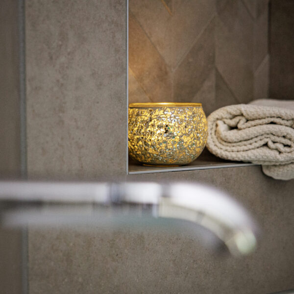 Wiederaufladbare LED-Teelichter mit Fernbedienung und bis zu 400 Stunden Brenndauer verbreiten ein warmes Licht auf einem Regal neben zwei gefalteten cremefarbenen Handtüchern vor einer grau gekachelten Wand.