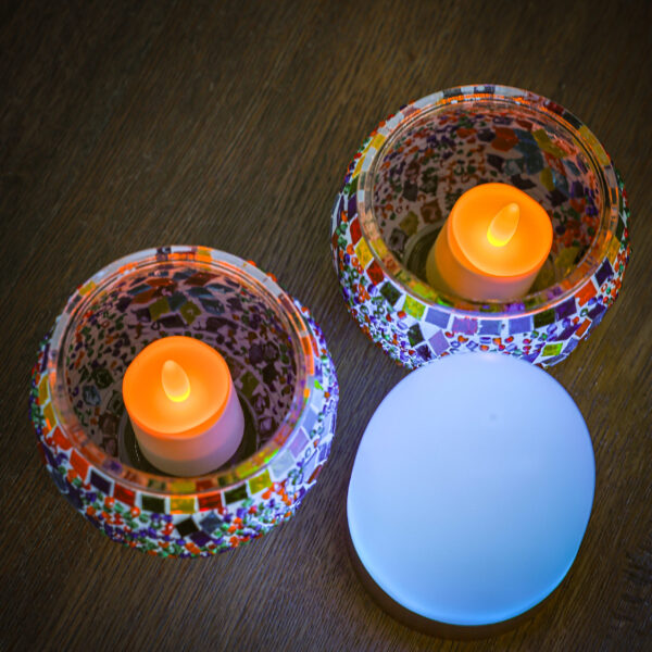 Zwei wiederaufladbare LED-Teelichter in Warmweiß leuchten in Mosaik-Glashaltern auf einer Holzoberfläche, neben einer ovalen weißen Lampe. Jedes wiederaufladbare LED-Teelicht hält bis zu 400 Stunden und ist wasserdicht; inklusive Fernbedienung (12er-Pack).