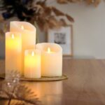 Eine wiederaufladbare LED-Wachskerze Ø 7,5 × 15 cm in Warmweiß steht auf einem Holztablett, umgeben von Dekoblättern und einem unscharfen gerahmten Foto. Die Szene wirkt einladend und gemütlich.