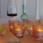 Eine Nahaufnahme von warmweißen, wasserfesten, wiederaufladbaren LED-Teelichtern (12er-Pack, bis zu 400 Stunden Brenndauer, mit Fernbedienung) auf einem Holztisch, neben einem Weinglas, grünen Trauben und einem türkisfarbenen Kerzenständer im Hintergrund.