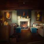 Ein gemütliches Wohnzimmer mit beigen Sofas, zwei blauen Hockern, einem beleuchteten Kamin und einer wiederaufladbaren LED-Wachskerze Ø 7,5 × 15 cm - warmweiß, die warmes Licht spendet. Große Porträts flankieren den Kaminsims unter einem montierten Hirschkopf und sorgen für eine einladende Atmosphäre.