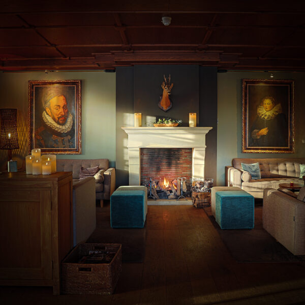 Ein gemütliches Wohnzimmer mit beigen Sofas, zwei blauen Hockern, einem beleuchteten Kamin und einer wiederaufladbaren LED-Wachskerze Ø 7,5 × 15 cm - warmweiß, die warmes Licht spendet. Große Porträts flankieren den Kaminsims unter einem montierten Hirschkopf und sorgen für eine einladende Atmosphäre.